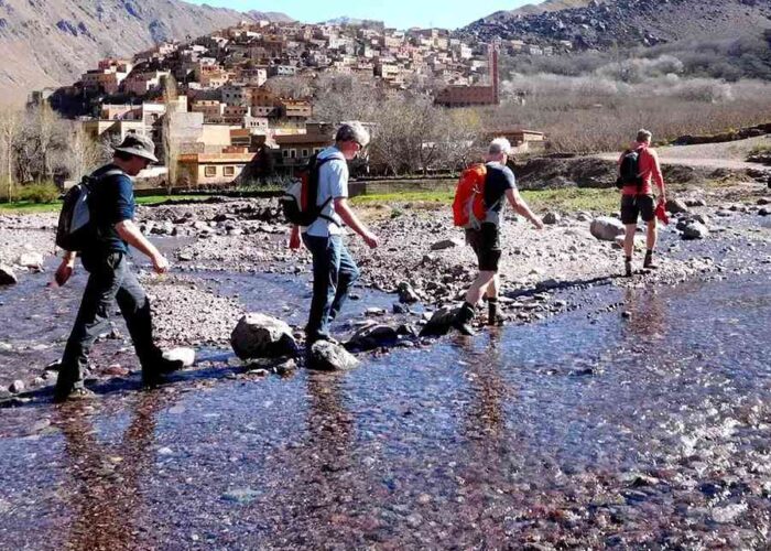 Trekkers crossing a rocky stream with a traditional Berber village in the backdrop in the Atlas Mountains.