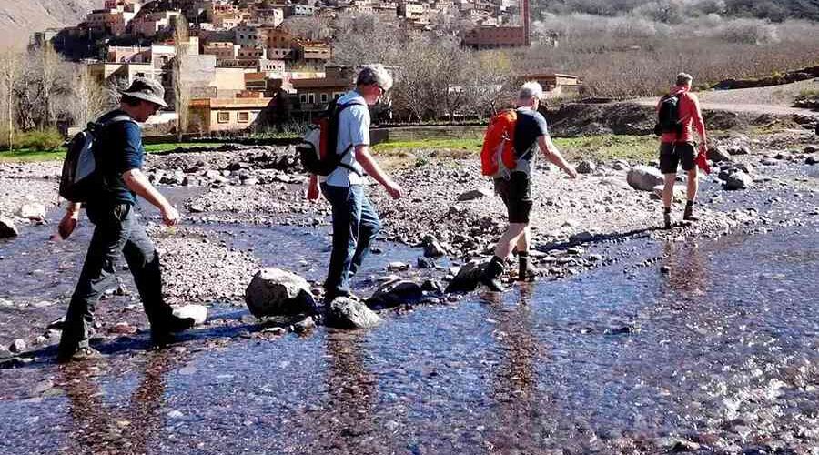 Trekkers crossing a rocky stream with a traditional Berber village in the backdrop in the Atlas Mountains.