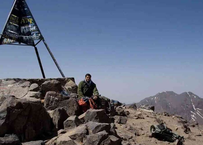 Hiker at Mount Toubkal summit with triangular sign.