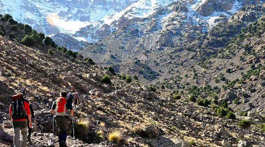Hikers on a rocky trail with the snow-dusted peaks of Mount Toubkal in the distance.