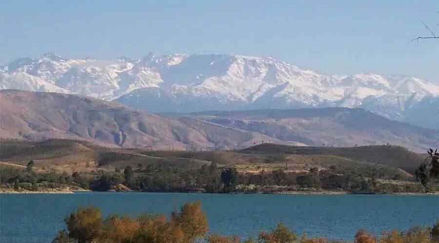 Tranquil lake in the foreground with the snow-capped Atlas Mountains towering in the background, under a clear blue sky.