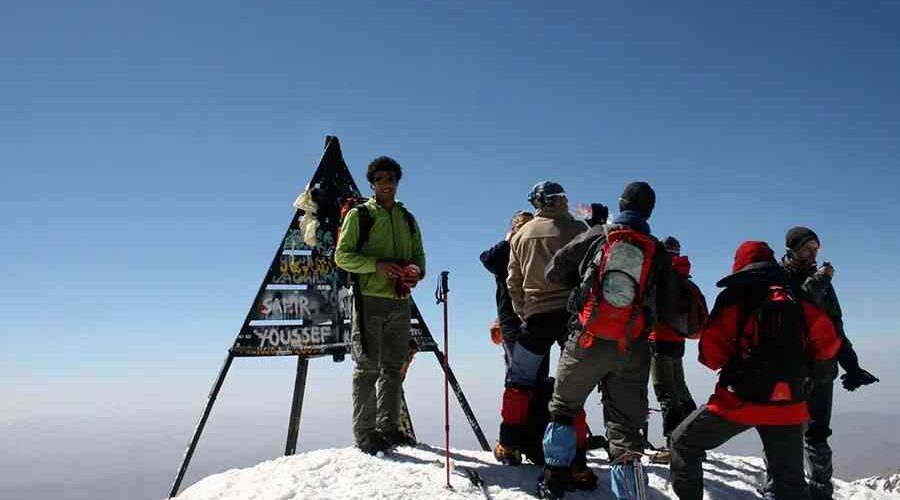 Group of hikers gathered around the summit sign of Mount Toubkal, the highest peak in the Atlas Mountains.