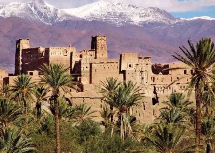 Historic Kasbah with snow-capped Mount Toubkal in the background, surrounded by lush palm trees under a clear blue sky in Morocco.