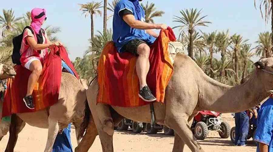 Tourists on a camel ride in Marrakech, dressed in traditional scarves, with quad bikes and palm trees in the background.