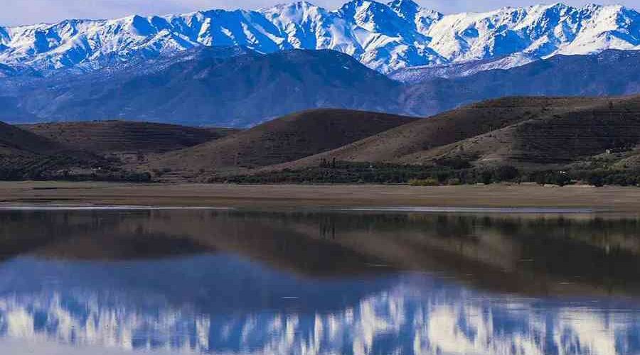 Mirror-like lake reflecting snow-capped mountains under a clear blue sky in Agafay Desert.