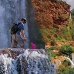 Two hikers exploring the Ouzoud Waterfalls with mist rising from the cascading waters and lush greenery in the background.