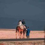 Two travelers on a camel in Agafay Desert with guide walking alongside.