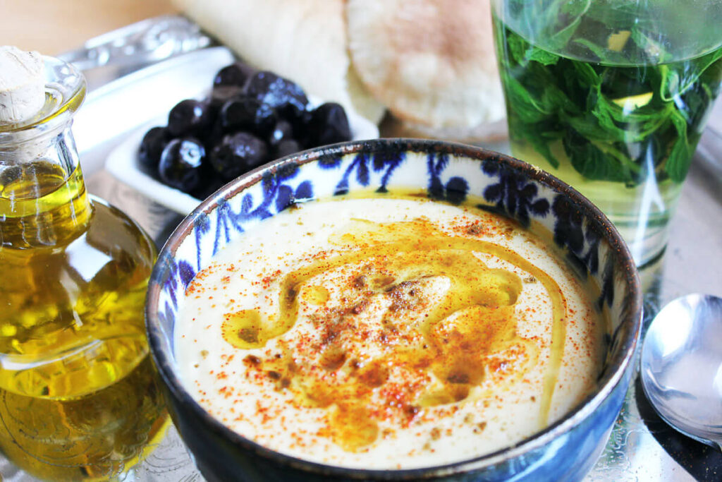 Creamy Moroccan Bissara soup in a traditional blue patterned bowl, drizzled with olive oil and sprinkled with spices, with fresh herbs and olives in the background.