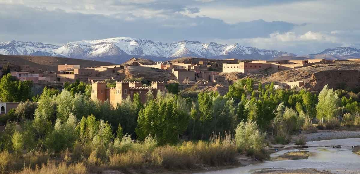 A panoramic view of the Dades Valley with traditional Moroccan architecture and lush greenery, against the backdrop of the snow-dusted Atlas Mountains.