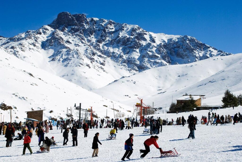 Crowds enjoying a sunny day at Oukaïmeden ski resort in the Moroccan Atlas Mountains, with snow-covered slopes and ski lifts in the background.