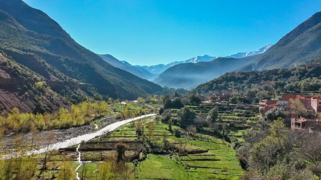 Verdant Ourika Valley with a flowing river and the snow-capped High Atlas Mountains in the distance.