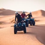 Group of adventurers riding quad bikes across the smooth sand dunes of Morocco Sahara Desert at dusk.