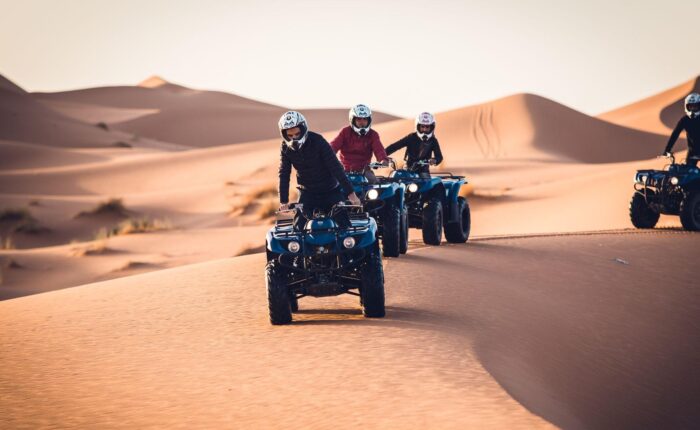 Group of adventurers riding quad bikes across the smooth sand dunes of Morocco Sahara Desert at dusk.