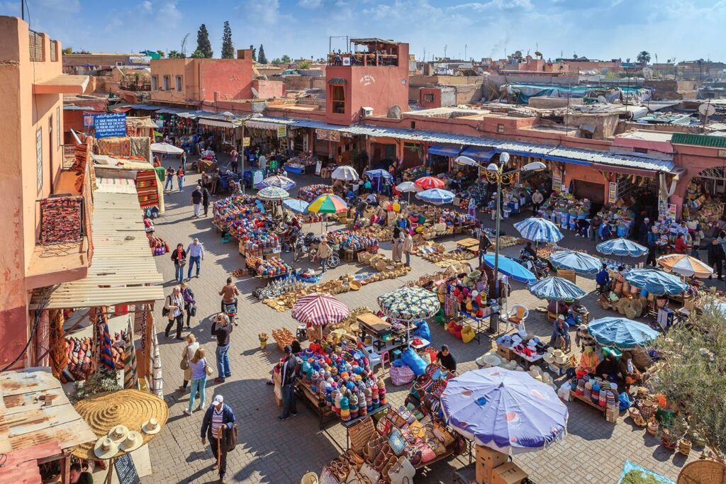 Overhead view of a bustling Rahba Kedima Square in Marrakech, with colorful market umbrellas and diverse local crafts on display.