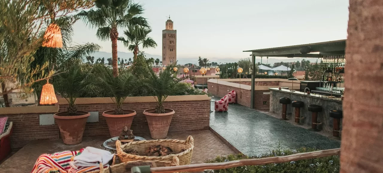 Cozy rooftop terrace of a Marrakech restaurant with views of Koutoubia Mosque amid palm trees at dusk.