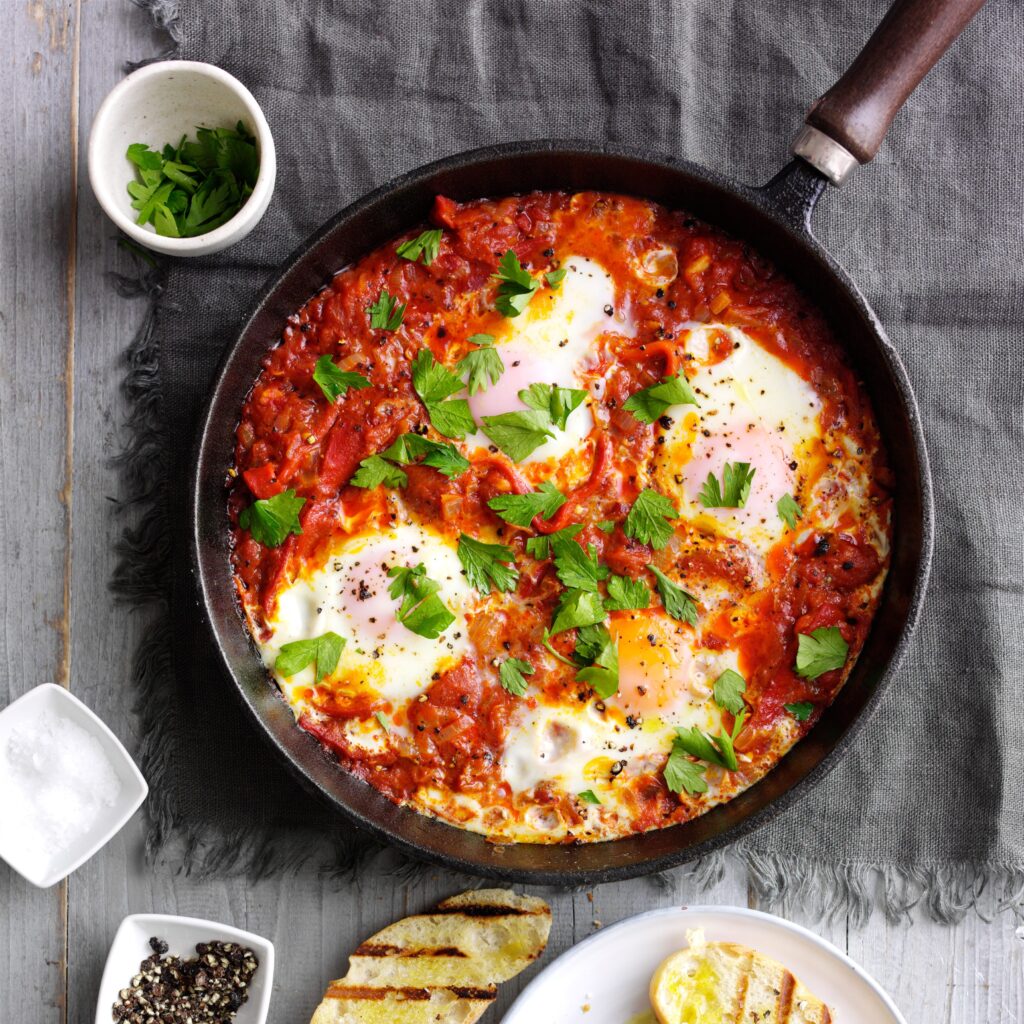 A skillet of Shakshuka with poached eggs in a rich tomato sauce, garnished with fresh parsley, served alongside crusty bread.