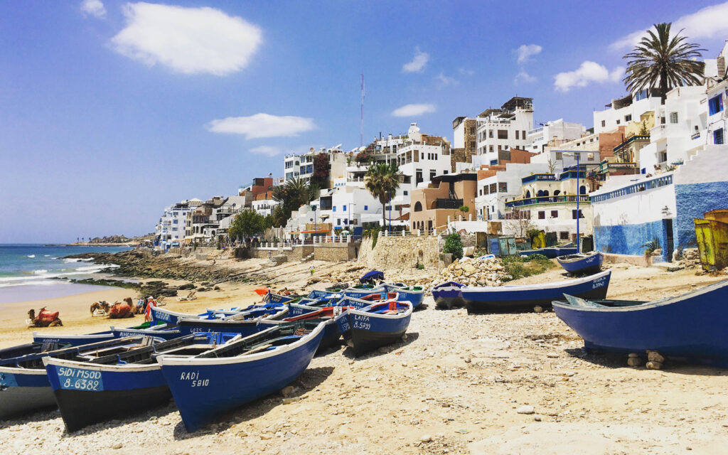 Blue fishing boats on the sandy shore of Taghazout beach with white and blue buildings in the background, under a sunny Moroccan sky.