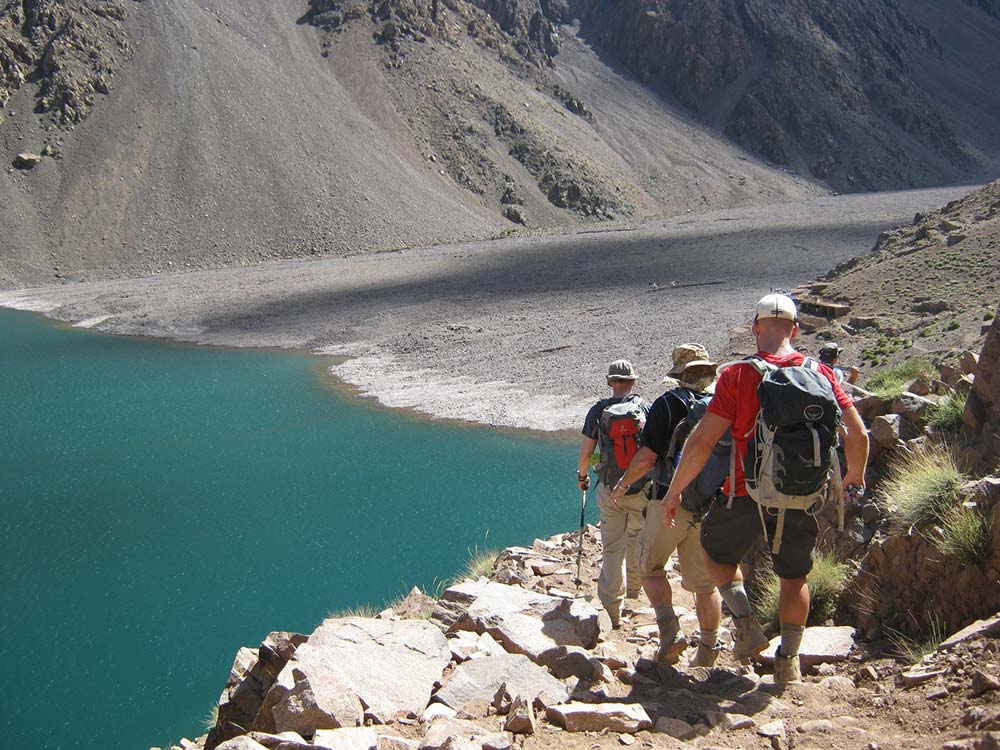 Trekking enthusiasts descending a trail alongside the teal waters of Ifni Lake in the Atlas Mountains.