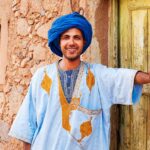 Smiling Moroccan man in traditional blue djellaba and turban standing by an old door in a Kasbah.