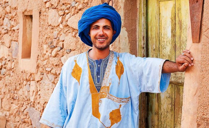Smiling Moroccan man in traditional blue djellaba and turban standing by an old door in a Kasbah.