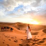 Woman in traditional attire watching the sunset over the Sahara dunes with a camel caravan in the background