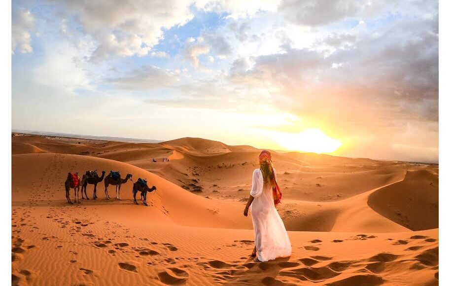 Woman in traditional attire watching the sunset over the Sahara dunes with a camel caravan in the background