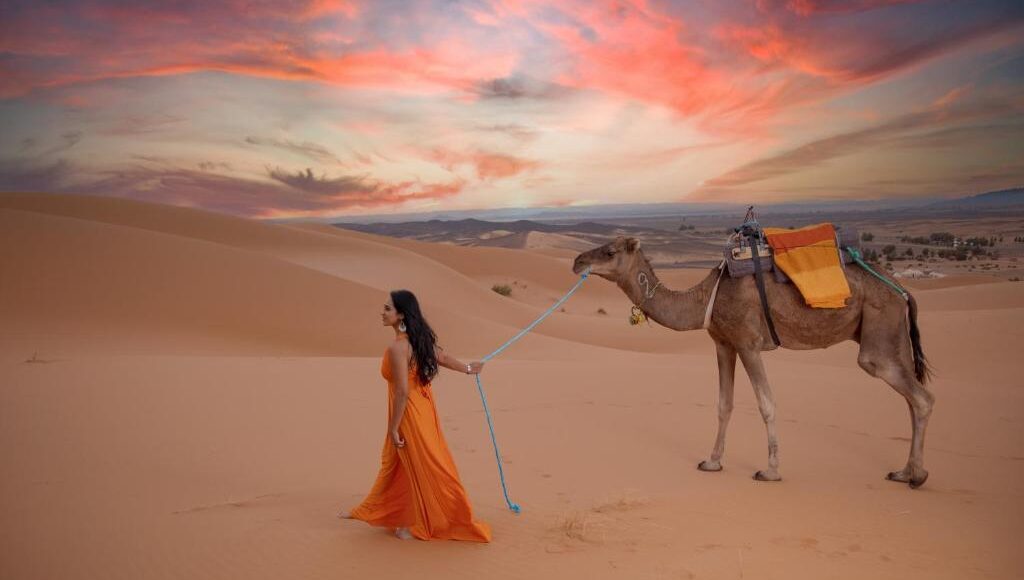 Woman in orange dress leading camel at sunset in Merzouga dunes.