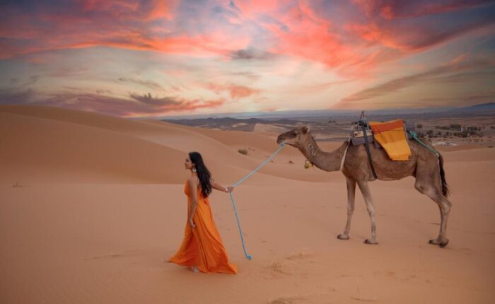 Woman in orange dress leading camel at sunset in Merzouga dunes.