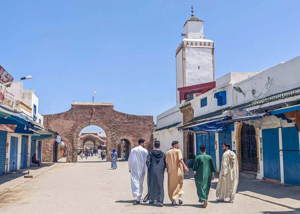Local residents walking towards a mosque under the clear blue sky in a traditional Moroccan market street during Ramadan.