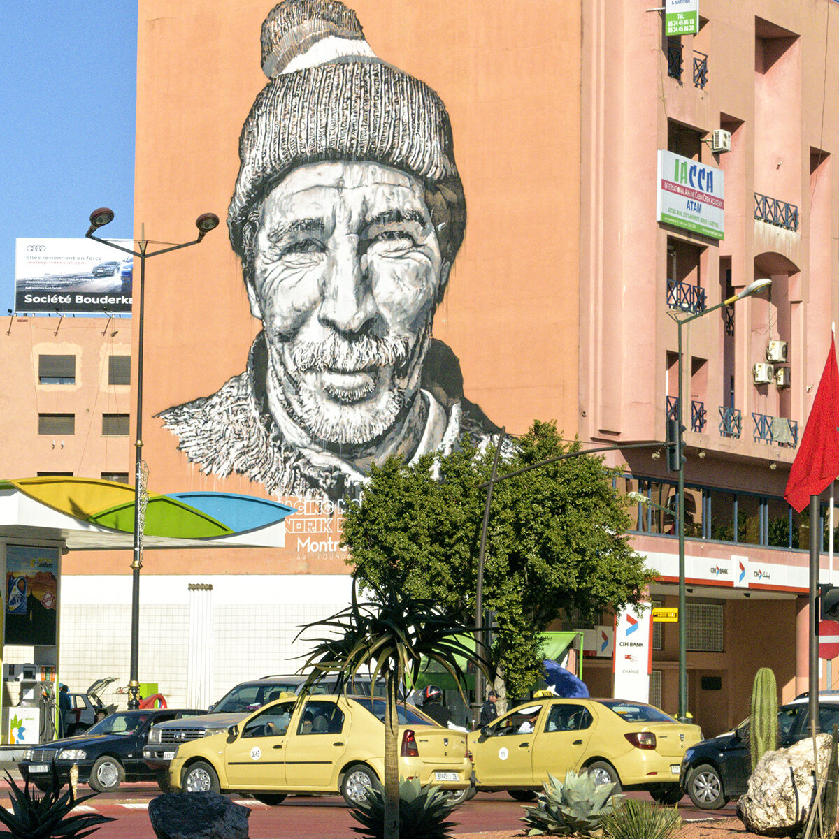 Giant mural of a local man's face on a building in Marrakech, with taxis and palms in the foreground.