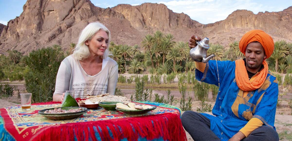 A woman tourist enjoying a traditional Moroccan tea served by a man in a blue turban outdoors with mountains and greenery in the background.
