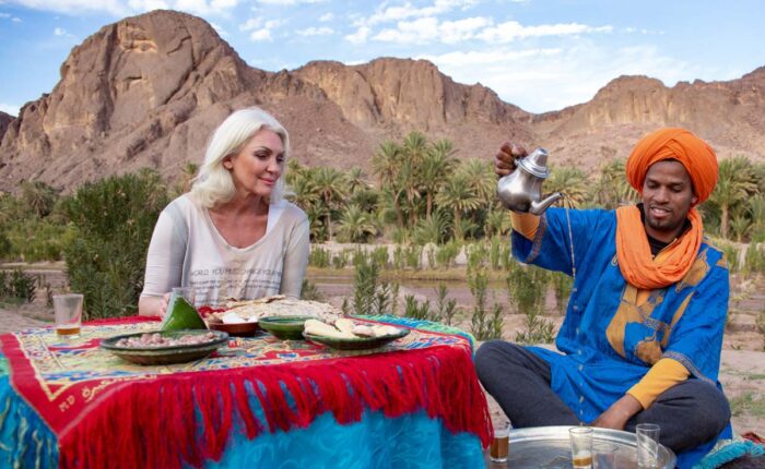 A woman tourist enjoying a traditional Moroccan tea served by a man in a blue turban outdoors with mountains and greenery in the background.