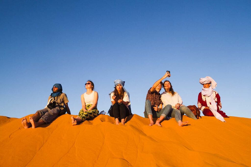Group of tourists seated on a golden sand dune in the Moroccan Sahara Desert, enjoying the serene sunset during Ramadan.