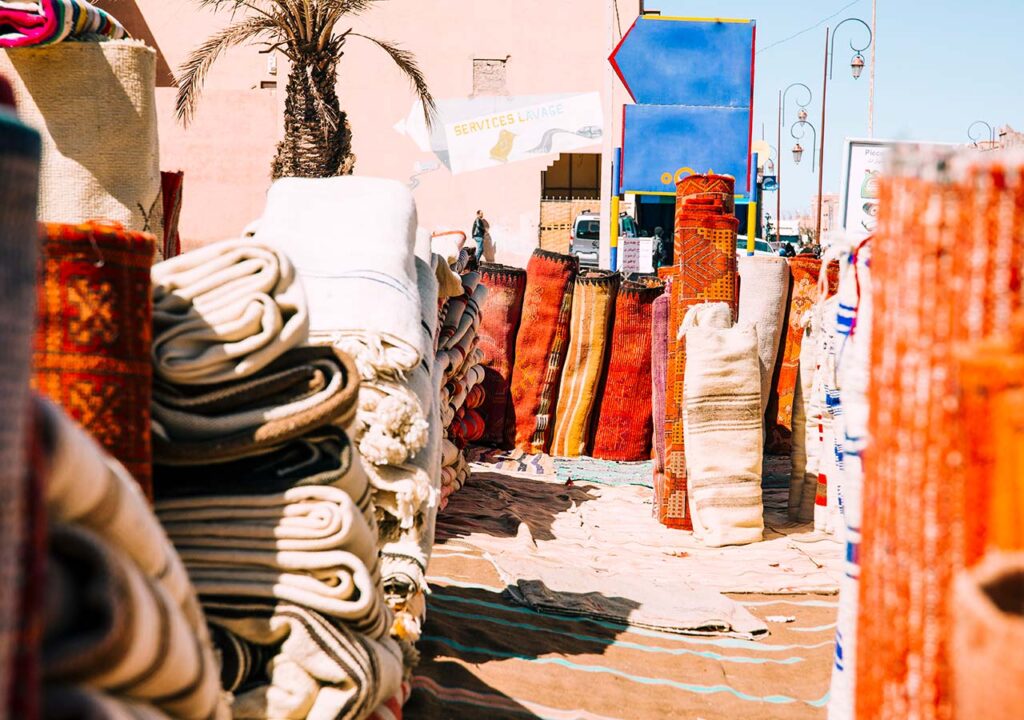 Colorful Moroccan rugs displayed in an open-air market with palm trees and blue skies in the background during Ramadan.