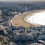Aerial view of Agadir's expansive sandy beach and curved bay, with palm-lined boulevards and white buildings, during a day trip from Marrakech.