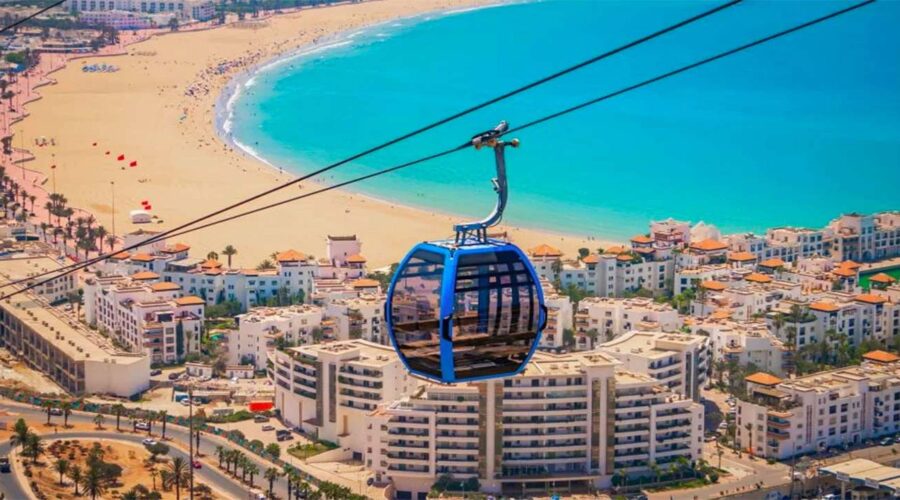 Vibrant cable car with panoramic views gliding over the bustling city and the sweeping curve of Agadir's azure beachfront, capturing the essence of coastal Morocco.