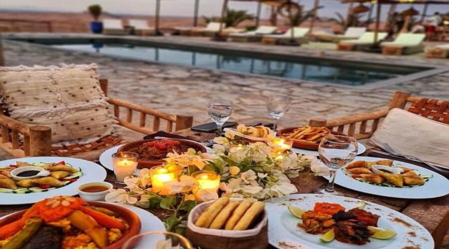 An exquisite Moroccan dinner spread by a poolside in the Agafay Desert, featuring couscous, tagines, and an assortment of appetizers, illuminated by candlelight at dusk.