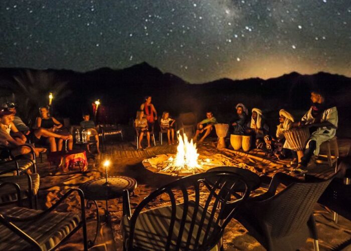 Group of people seated around a roaring bonfire under a starry sky in the Agafay Desert during a sunset camel ride event, with traditional Moroccan lamps and drums.