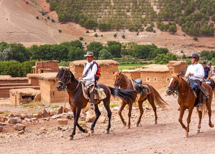 A group of riders on horseback led by a guide in traditional attire, traversing a dusty trail past earthen buildings with the lush Atlas Mountains in the backdrop.