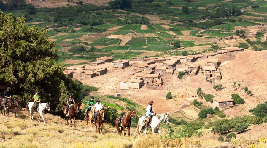 Horseback riders meander down a hillside trail overlooking a traditional Moroccan village amidst the verdant fields of the Atlas Mountains.