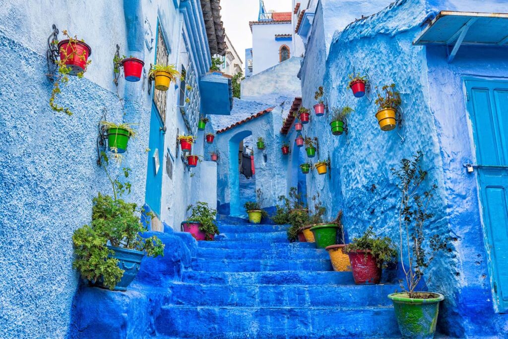 Colorful flower pots in red, yellow, and green hanging on the blue-washed walls of a narrow stairway in Chefchaouen, Morocco, with blue doors and a small arched entryway adding to the picturesque scene.