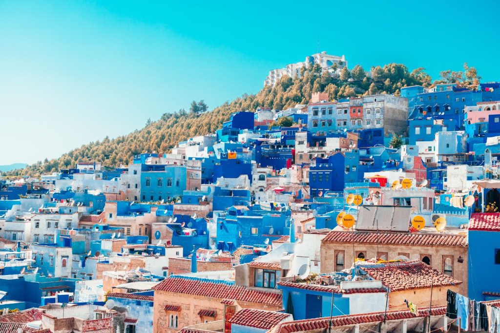 A vibrant view of Chefchaouen, Morocco's Blue City, showcasing the densely packed blue houses with terracotta roofs set against the green hills and clear blue sky, with satellite dishes dotting some rooftops.