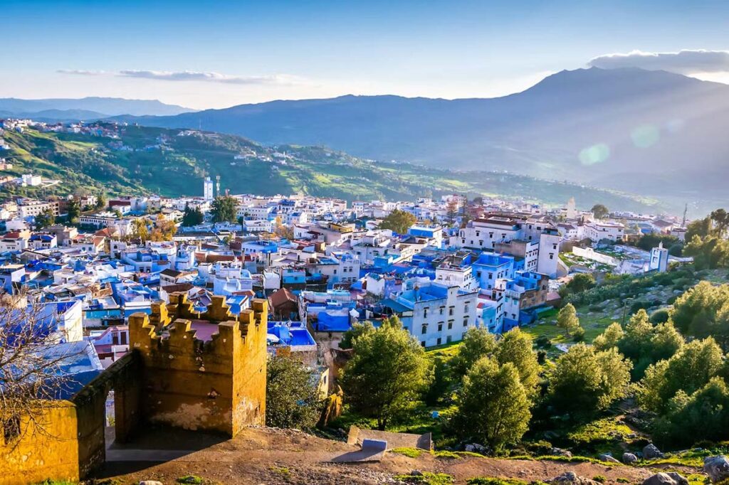 Elevated view of Chefchaouen at sunset, showcasing the city's iconic blue-painted houses and winding streets, with the surrounding lush green hills and a backdrop of majestic mountains under a soft golden sky.