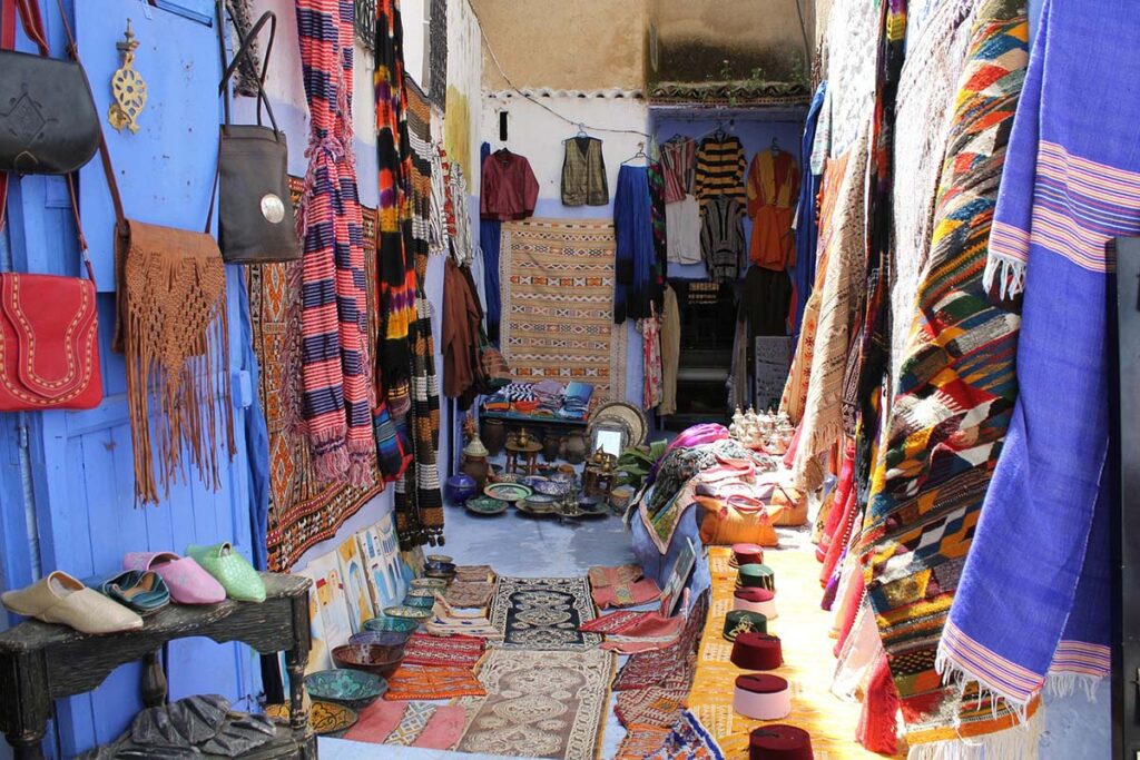 Colorful Moroccan market stall lined with traditional goods: vibrant woven rugs, leather bags, clothing, and footwear against a blue-painted wall, reflecting Chefchaouen's rich artisanal culture.