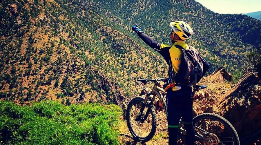 Mountain biker in yellow helmet stands pointing towards the lush greenery and rolling hills of the Atlas Mountains, with a bike beside them on the trail.