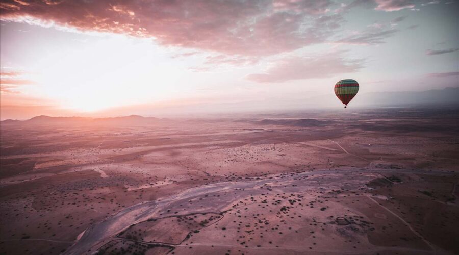 Hot air balloon with colorful stripes soaring over the serene desert landscape near Marrakech at sunrise, casting a shadow on the sandy ground below.