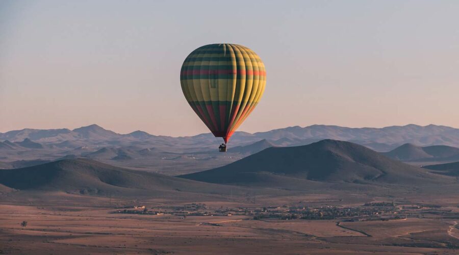 A striped hot air balloon floats over the rolling hills near Marrakech in the golden light of early morning.