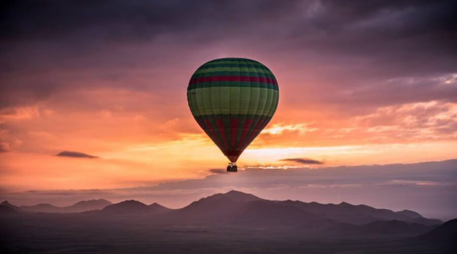 Colorful hot air balloon against a dramatic sunrise over the Marrakech horizon, with silhouetted mountains in the background.