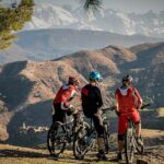 Three mountain bikers stand with their bikes on a hill overlooking the Atlas Mountains and Berber villages in Morocco, with snow-capped peaks in the distance.