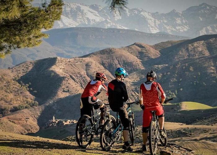 Three mountain bikers stand with their bikes on a hill overlooking the Atlas Mountains and Berber villages in Morocco, with snow-capped peaks in the distance.